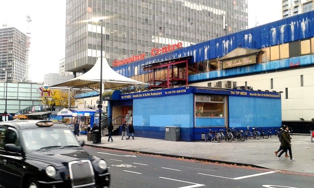 Elephant and Castle Shopping Centre’s front exterior, showcasing its urban design against a cloudy sky. London Black Cab in the bottom left of the frame.
