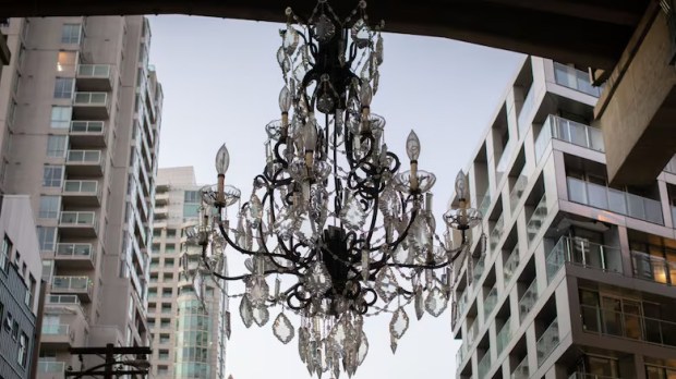 A large, illuminated chandelier installation spins slowly beneath Vancouver’s Granville Bridge at night. Below it, the underpass is dark and largely empty — a site where unhoused people often sleep.