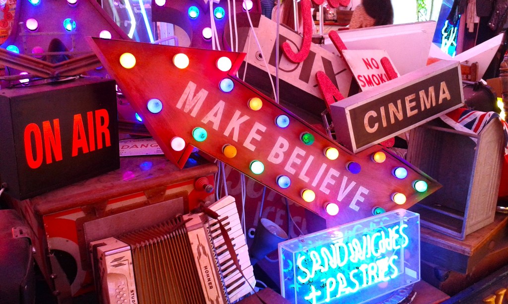 A colorful, chaotic collection of neon signs and vintage objects piled together in God’s Own Junkyard, Walthamstow. Visible signs include a glowing “On Air” light, a double-sided arrow reading “Make Believe” surrounded by bright bulbs, a “Cinema” sign, and a weathered Hohner accordion. Various other illuminated signs and retro items create a vibrant, nostalgic collage of light and culture.